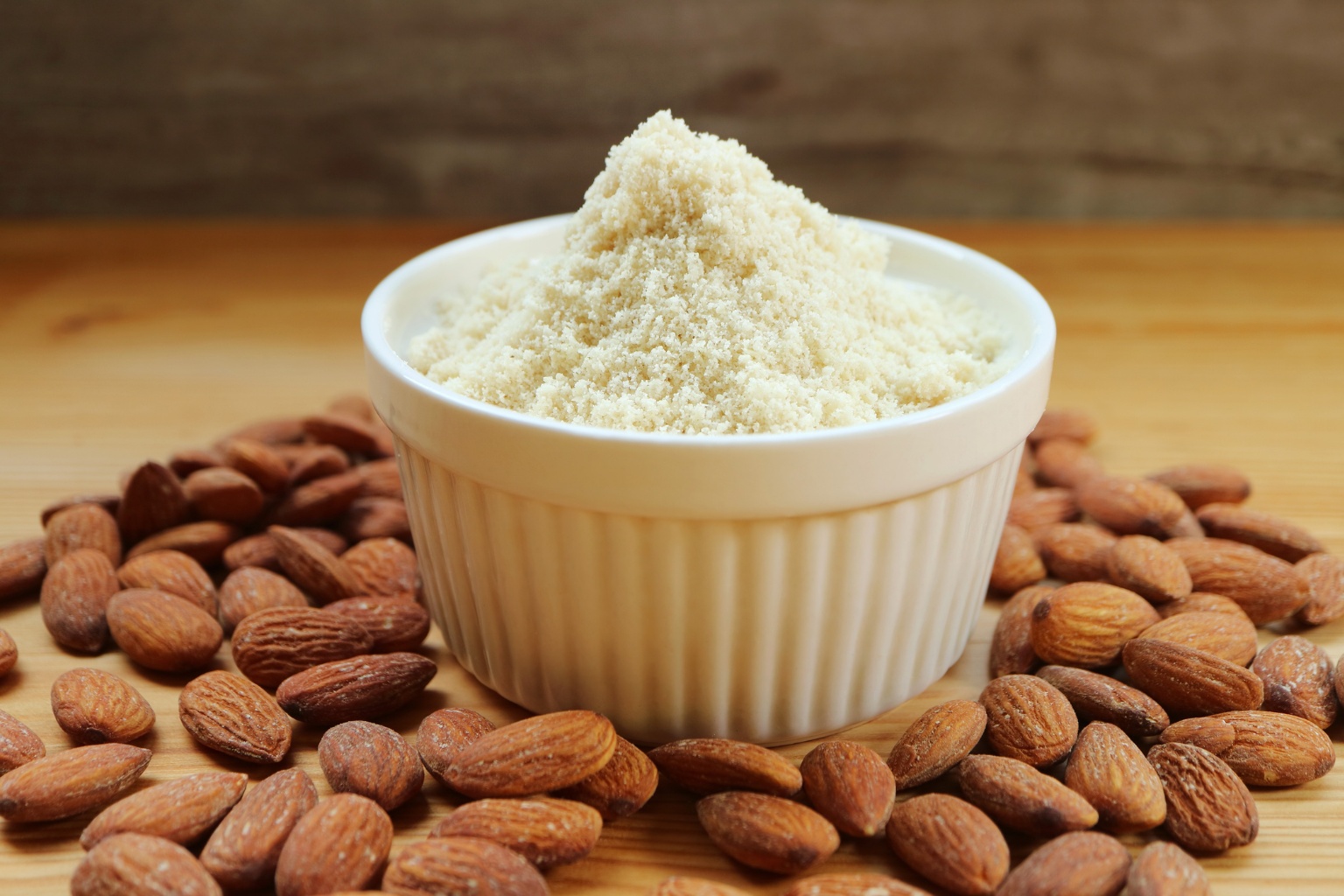 Mąka migdałowa w jasnym naczyniu oraz migdały rozrzucone po stole.